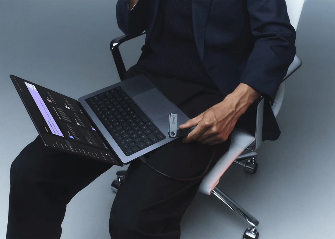 A Ledger Nano S Plus plugged into the side of a laptop by its USB cable while the owner holds the laptop on their lap