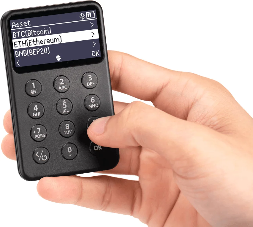 A hand holding the SafePal X1 hardware wallet, showing the 12-button keypad and the asset list on screen