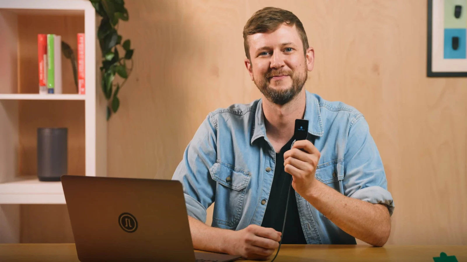 Trezor Safe 3 held up to the camera showing its monochrome OLED screen, with a laptop on the desk