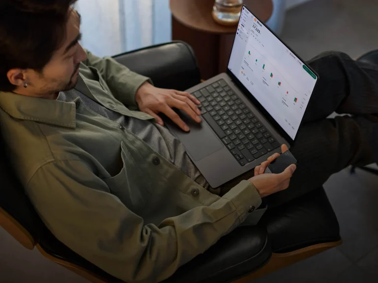 A person holding a Trezor Safe 7 hardware wallet next to a laptop running the Trezor Suite app