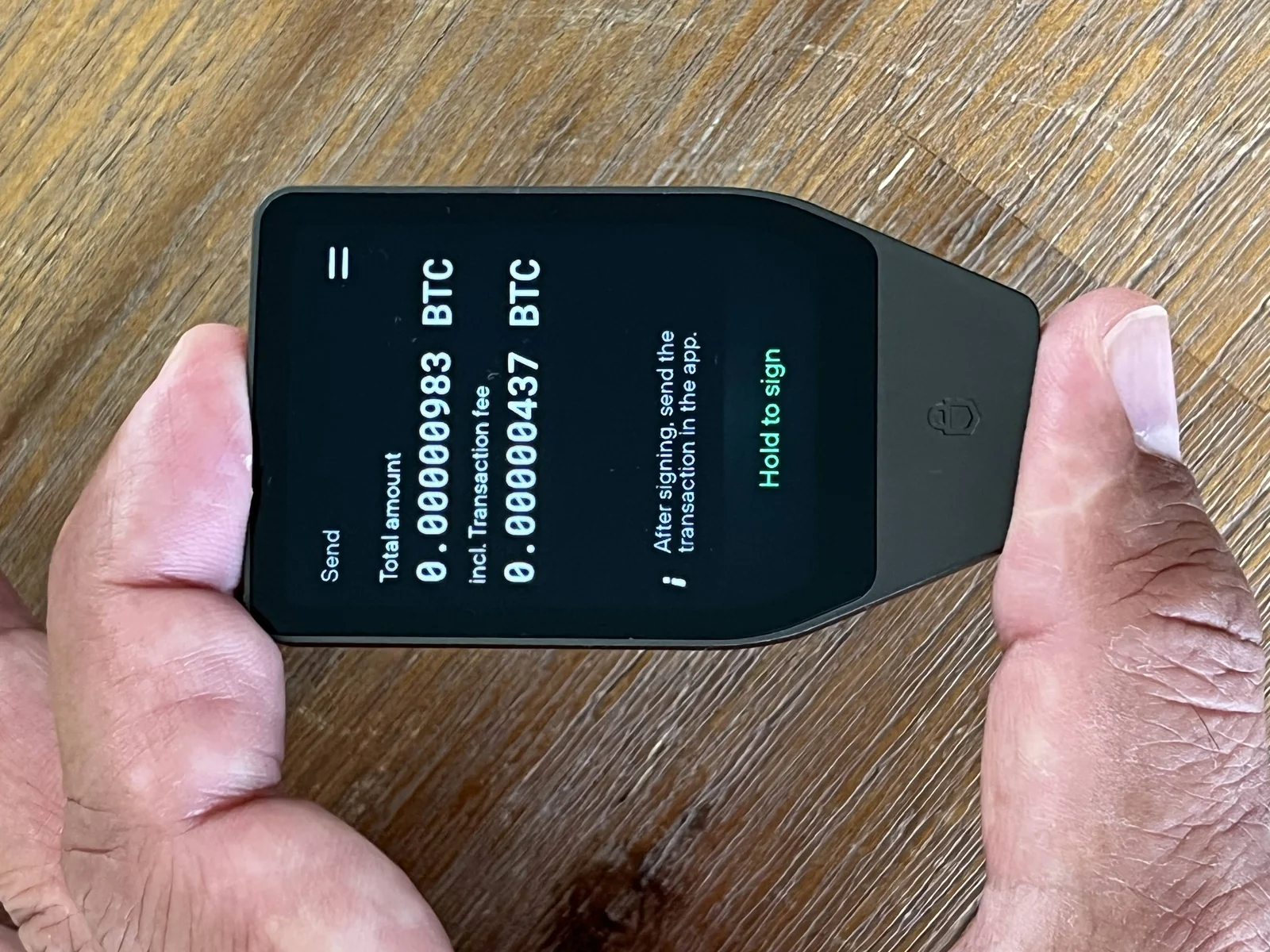 A Trezor Safe 7 hardware wallet held in both hands showing a hold to sign confirmation screen, the same screen used to confirm a passphrase